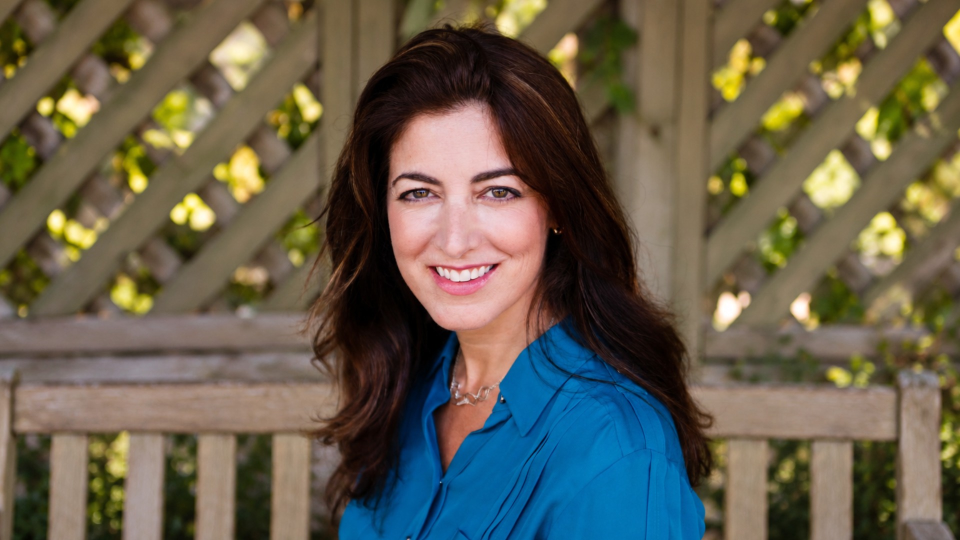 A smiling woman with long dark hair wearing a blue blouse sits on a wooden bench outdoors, with a leafy lattice in the background.